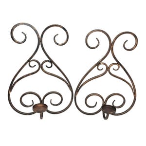 May include: Two ornate, bronze-colored metal wall sconces. Each sconce features a heart-shaped design with decorative swirls and a small, circular candle holder. The sconces are designed to hold candles and add a touch of elegance to any room.