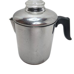 Pode incluir: Uma cafeteira percoladora de aço inoxidável para fogão com acabamento metálico polido, alça preta resistente ao calor e botão de vidro transparente na tampa. O corpo cilíndrico inclui um pequeno bico para servir na lateral.
