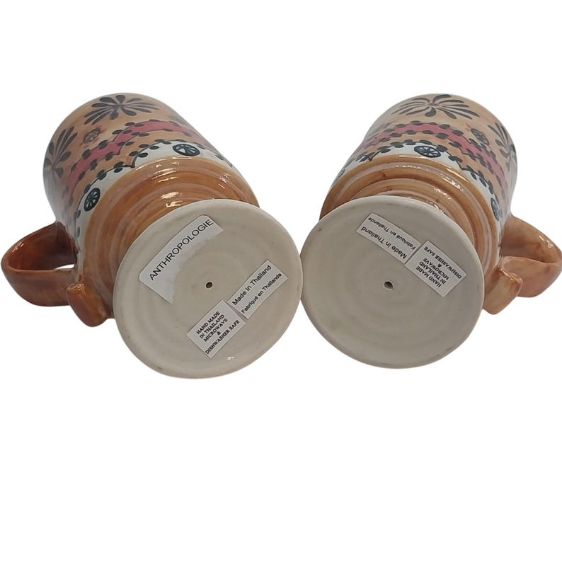 Op de afbeelding: Twee keramische mokken met handvatten, met een bruinachtige basis en een decoratief patroon van roze, zwarte en witte bloemmotieven. De mokken hebben een brede basis en zijn voorzien van de labels "ANTHROPOLOGIE" en "Made in Thailand".