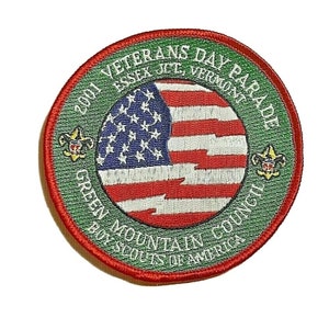 Peut inclure: Un écusson brodé vert et rouge avec le texte "2001 Veterans Day Parade Essex Jct, Vermont Green Mountain Council Boy Scouts of America". L'écusson présente un drapeau américain stylisé au centre.