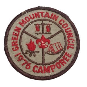 May include: A brown and tan embroidered patch with the text "Green Mountain Council 1976 Camporee" in red and brown lettering. The patch features a campfire, a tent, a fleur-de-lis, and two buckets hanging from a hook.