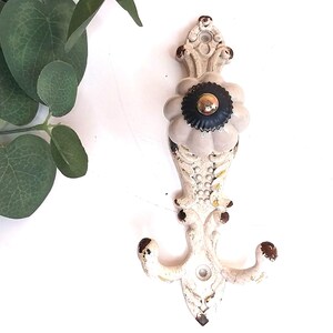 May include: A wall-mounted hook featuring an off-white, distressed metal backplate with ornate scrollwork and brown paint chips. A large, round, light grey ceramic knob with a dark brown, fluted centre and a metallic gold-toned top sits at the centre.
