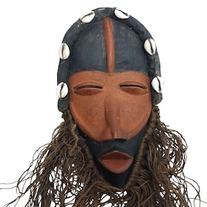 May include: A carved wooden mask with a black and brown color scheme. The mask features a prominent nose, closed eyes, and a mouth. White seashells adorn the top of the mask. Brown, rope-like material hangs from the bottom.