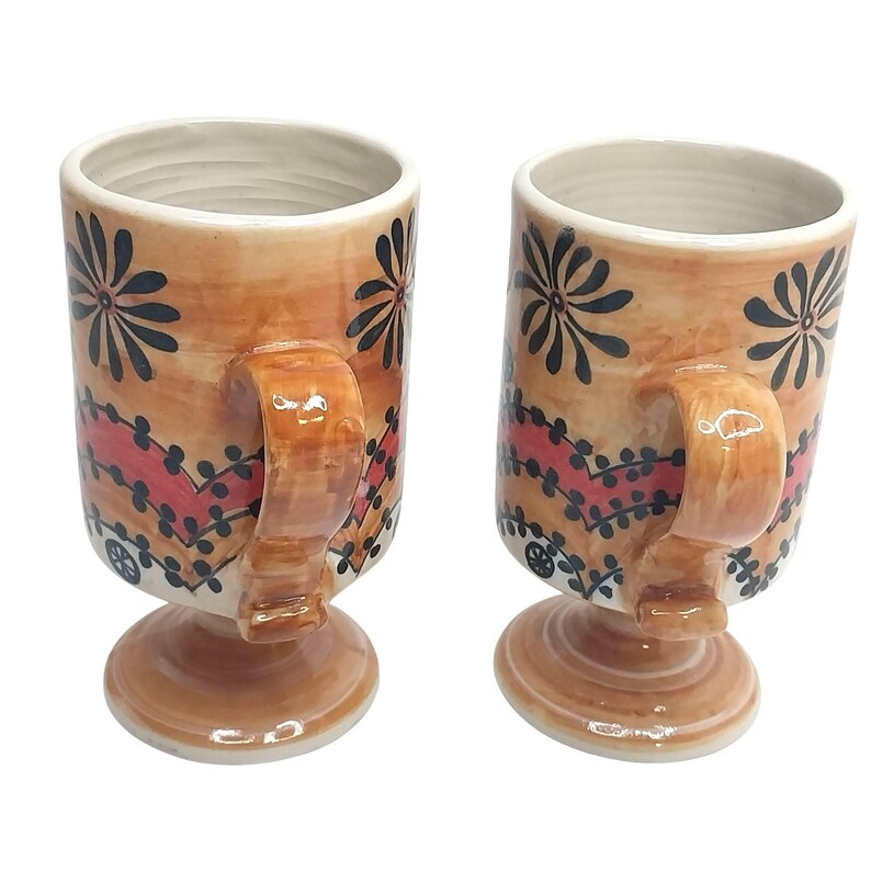 Op de afbeelding: Twee keramische mokken met een bruine basis en handvat, met een bloemenontwerp in zwart en rood. De mokken hebben een voetstuk en een cr&egrave;mekleurig interieur. Het ontwerp omvat zwarte bloemen en een rood-zwart patroon.