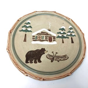 May include: A round ceramic plate with a speckled beige surface and a brown, textured rim. The center features a painted scene of a log cabin with snow on the roof, three green pine trees with white snow, a brown bear, and a canoe with two paddles.