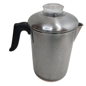 May include: A stainless steel percolator coffee pot featuring a polished metal body, a black heat-resistant handle, a matching metal lid, and a clear glass knob on top. The coffee pot has a tapered spout on the side and a copper-toned base.
