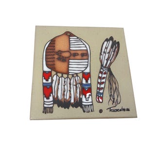 May include: Square ceramic tile with a Native American-inspired design. It features a central motif of a shield and feathers, with red, white, and blue accents. The tile has a beige background and is signed "Teissedre 86".