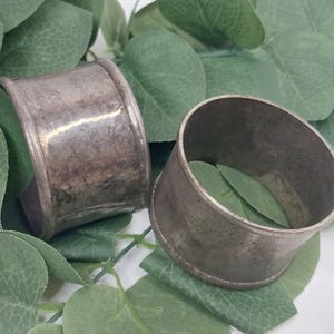 Op de afbeelding: Twee antieke zilveren servetringen met een verweerde uitstraling. De ringen zijn cilindrisch met een licht uitlopend ontwerp. De achtergrond toont groene bladeren, wat een natuurlijk contrast geeft met de metalen afwerking. Voor formele diners.