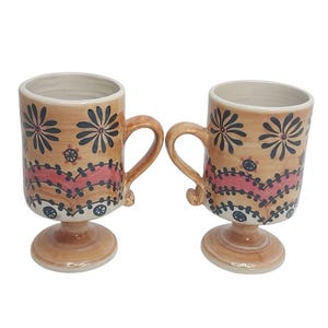 Op de afbeelding: Twee keramische mokken met handvatten, met een bruinachtige basiskleur en ingewikkelde bloemmotieven in zwart en rood. De mokken hebben een voet en zijn geschikt voor warme dranken.
