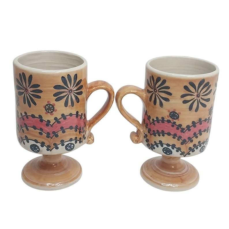 Op de afbeelding: Twee keramische mokken met handvatten, met een bruinachtige basiskleur en ingewikkelde bloemmotieven in zwart en rood. De mokken hebben een voet en zijn geschikt voor warme dranken.