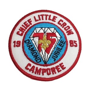 Può includere: Una toppa ricamata bianca e rossa con il testo "Chief Little Crow 1919 Diamond Jubilee 1985 Camporee". Al centro della toppa c'è un diamante rosso con contorno bianco e un giglio dorato.