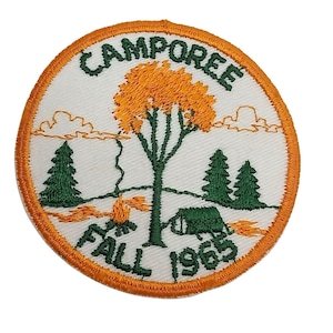 Może przedstawiać: Biała naszywka haftowana z pomarańczową obwódką. Naszywka przedstawia scenę kempingową z drzewem, ogniskiem, namiotem i dwoma sosnami. Tekst "CAMPOREE FALL 1969" jest wyszyty na zielono.