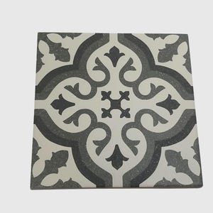 May include: Square decorative tile with a repeating floral pattern in shades of gray and white. The design features a central cross-like motif surrounded by swirling, stylized leaves and curves. The tile has a matte finish.