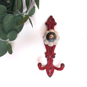 May include: A red metal wall hook featuring an ornate, scrollwork design with a central, cream-coloured, flower-shaped ceramic knob. The knob has a small, dark, circular centre with a metallic gold-toned accent at the very top of the piece.