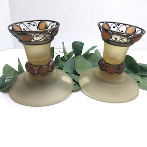 May include: A pair of candle holders featuring frosted pale yellow glass bases and stems. Each holder is accented with dark bronze metal filigree at the top and a ring of circular amber-colored glass inlays around the neck and rim.