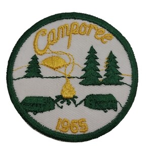 Może przedstawiać: Biała naszywka haftowana z zielonymi i żółtymi detalami. Naszywka przedstawia scenę z ogniskiem z dwoma drzewami, ogniskiem i dwoma namiotami. Tekst "Campore" i "1965" jest wyszyty na żółto.