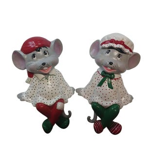 Puede incluir: Dos figuras de ratón grises con orejas grandes y caras sonrientes. El ratón de la izquierda lleva un gorro rojo y una camisa blanca de lunares con pantalones rojos. El de la derecha lleva un gorro blanco con borde rojo, camisa de lunares y lazo verde.