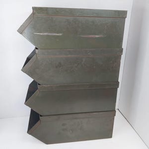 May include: A stack of four olive green metal storage bins with an open-front design. Each rectangular bin features a slanted front edge, a flat top, and a weathered finish with visible surface scratches and signs of wear on the metal exterior.