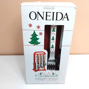 May include: A white Oneida stainless steel flatware set in a box labeled Holiday Colormates. The packaging features green Christmas trees and red snowflakes. The set includes 24 pieces for 6 people and a portable caddy, with utensils displaying a festive tree and snowflake pattern on the handles.