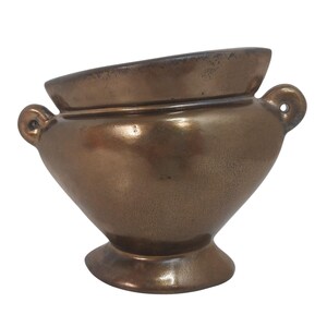 May include: A metallic bronze-colored ceramic vessel with a wide, slanted rim and a pedestal base. The container features two small, circular handles on opposite sides, each with a central hole, and a smooth, reflective finish across its surface.