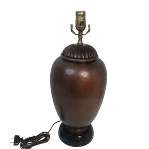 May include: A table lamp featuring a smooth, dark copper-toned urn-shaped body with a ribbed metal cap. The lamp sits on a circular black base and includes a brass-colored socket assembly at the top, accompanied by a brown electrical cord and plug.