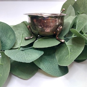 May include: A small, antique silver-colored bowl with three curved feet and a small spoon inside. The bowl is set on a bed of green eucalyptus leaves, creating a natural and elegant display. The bowl has a tarnished appearance.