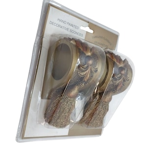 Op de afbeelding: Handgeschilderde decoratieve wandkandelaars in een doorzichtige plastic verpakking. De wandkandelaars zijn goud en bruin met een kwastjesontwerp. De verpakking leest "Timeless Classics Designed By".