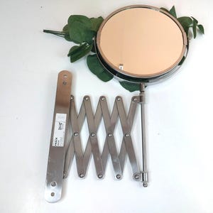 May include: A stainless steel wall-mounted mirror with a circular reflective surface and an extendable accordion-style arm. The base plate features a white label with the text IKEA FRÄCK 20325. The mirror is positioned near green leaves.