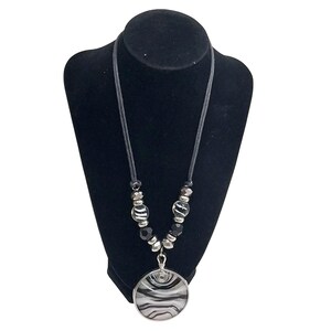 May include: A black and white pendant necklace displayed on a black mannequin. The necklace features a round pendant with a swirling pattern, black and silver beads, and a gray cord. The jewelry is designed for everyday wear.