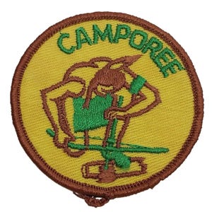 Może przedstawiać: Naszywka haftowana w kolorze brązowym i żółtym z napisem "CAMPOREE" w zielonych literach. Naszywka przedstawia kreskówkową ilustrację osoby trzymającej kubek i siedzącej na stołku.