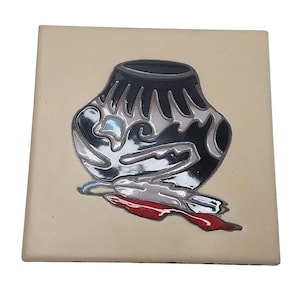Puede incluir: Una baldosa de cerámica cuadrada con fondo beige. La baldosa presenta un diseño negro y plateado de una olla nativa americana con una pluma roja y plateada. La olla tiene una forma única y estilizada.