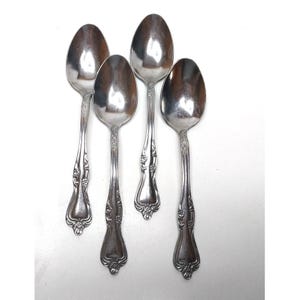 May include: A set of four silver-toned metal spoons arranged vertically. Each spoon features a polished oval bowl and a handle decorated with embossed floral and scrollwork patterns that extend from the neck down to the rounded base of the handle.