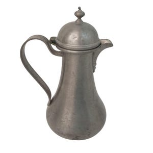 May include: A matte gray metal pitcher with a curved handle, a wide base, and a domed lid topped with a small spherical finial. The vessel features a short, pointed spout and a smooth, weathered surface texture throughout its entire exterior.