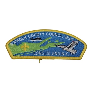 Könnte beinhalten: Ein gestickter Aufnäher in Blau und Grün mit dem Text "Suffolk County Council BSA Long Island N.Y.". Der Aufnäher zeigt ein stilisiertes Bild von Long Island, eine Möwe und eine Lilie.