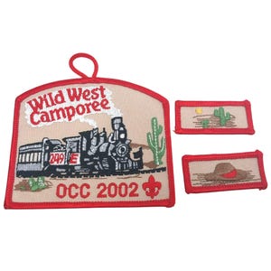 Może przedstawiać: Haftowane naszywki z motywem Dzikiego Zachodu. Główna naszywka przedstawia pociąg, kaktus i napis "Wild West Camporee OCC 2002". Dwie mniejsze naszywki przedstawiają pustynny krajobraz i kapelusz kowbojski.