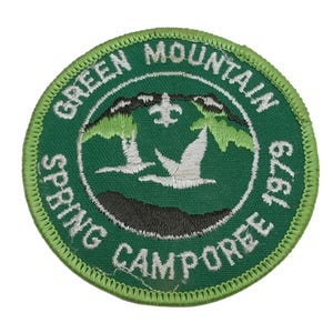 Może przedstawiać: Okrągła, zielona naszywka z haftem z napisem "GREEN MOUNTAIN SPRING CAMPOREE 1979". Naszywka przedstawia biały fleur-de-lis, dwa białe ptaki i zielone drzewa.