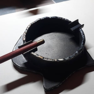 Puede incluir: Cenicero de metal negro con una superficie rugosa y texturizada. El cenicero tiene un borde elevado y una pequeña abertura rectangular en el lateral. Un palo de madera con una punta de latón descansa en el borde del cenicero.