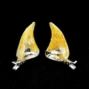 May include: A pair of gold glitter devil horn hair clips. The horns are curved, with a clear base and a sparkling gold glitter top. Each clip has a silver metal clip attached to the base.