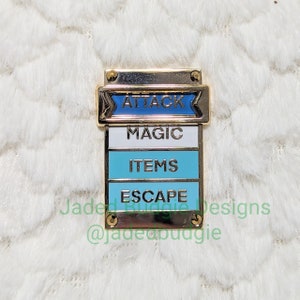 Puede incluir: Un pin de esmalte dorado con un diseño apilado. La sección superior dice "ATTACK" en blanco sobre una pancarta azul. Debajo hay secciones blancas, azules y verde azulado con las palabras "MAGIC", "ITEMS" y "ESCAPE".