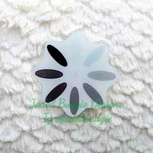 May include: A flower-shaped acrylic badge featuring a white base with eight oval petals in varying shades of black and gray, arranged in a radial pattern. The text Jaded Budgie Designs and Ig @jadedbudgie is printed in green across the center.