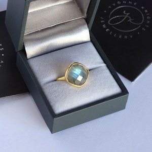 May include: A gold ring with a square-shaped, iridescent labradorite gemstone. The ring is in a black ring box with the text "Yvonne Knight Jewellery" on the lid.