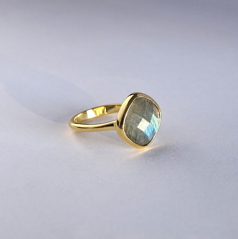 May include: A gold ring with a large, faceted, gray labradorite gemstone. The ring is set in a simple, modern style.
