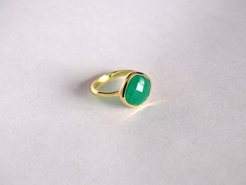 Puede incluir: Un anillo de oro con una piedra preciosa verde. La piedra preciosa es de forma cuadrada con bordes redondeados y tiene un corte facetado.