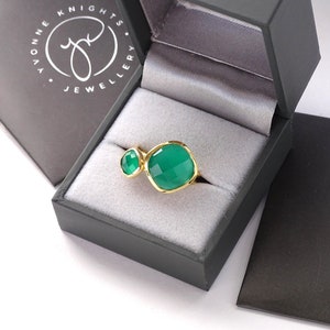 Puede incluir: Un anillo de oro con dos engastes de piedras preciosas verdes. El anillo est&aacute; en una caja de joyer&iacute;a negra con el texto "Yvonne Knights Jewellery" en la tapa.