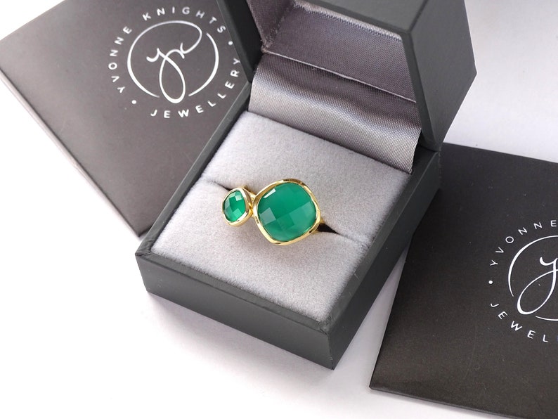 Puede incluir: Un anillo de oro con dos engastes de piedras preciosas verdes. El anillo est&aacute; en una caja de joyer&iacute;a negra con el texto "Yvonne Knights Jewellery" en la tapa.