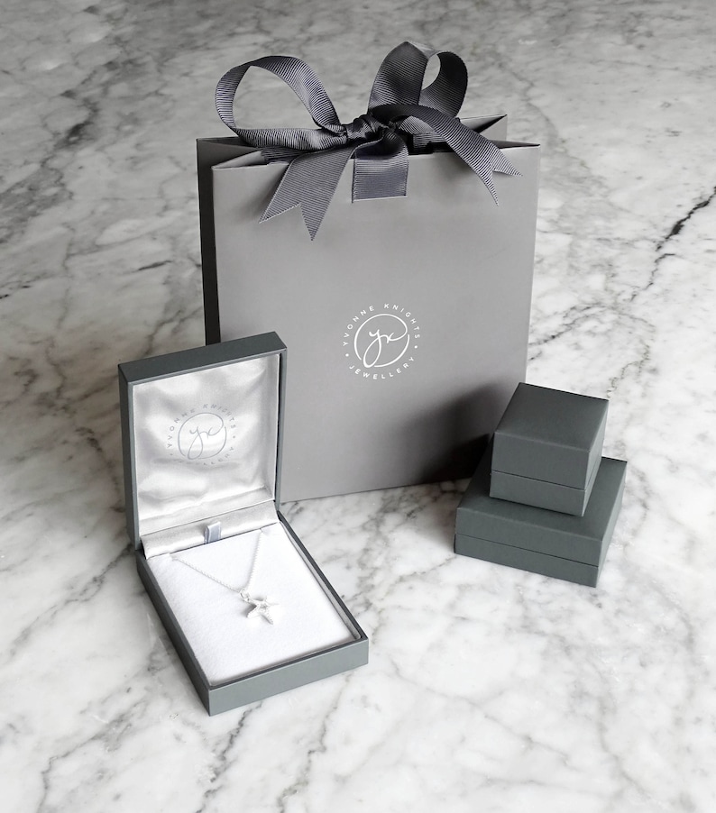 May include: A gray jewelry box with a silver necklace inside. The box has a logo that says "Yvonne Knight Jewellery". The necklace is in a gray gift bag with a gray ribbon.
