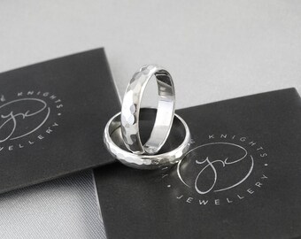 Peut inclure: Deux alliances en argent martelées empilées l'une sur l'autre. Les alliances sont sur une surface noire avec le texte "The Knights Jewellery" imprimé dessus.