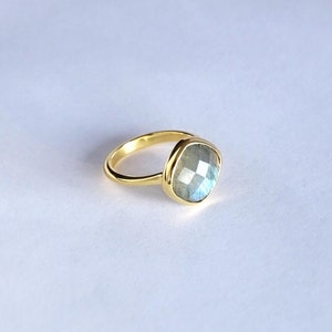 May include: A gold ring with a large, square-shaped labradorite gemstone. The gemstone has a blue-green iridescent sheen.