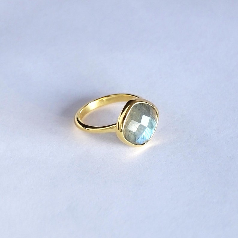 May include: A gold ring with a large, square-shaped labradorite gemstone. The gemstone has a blue-green iridescent sheen.