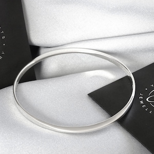 Könnte beinhalten: Ein silbernes Armreif-Armband auf einem weißen Stoffhintergrund. Das Armband ist schlicht und elegant mit polierter Oberfläche.